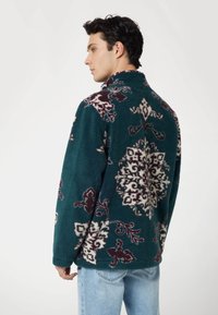 Tamsiai žalia pūkinė striukė su aukštu apykakle, puošta balta ir bordo gėlių raštu. Matoma užtrauktuko detalė apačioje, laisvas fasonas.