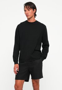 Man in een eenvoudig zwart langemouwen shirt en zwarte shorts, staande met één hand in de zak tegen een witte achtergrond.