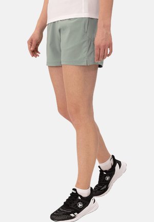 Person standing, wearing light green athletic shorts, white shirt, white socks, and black running shoes against a plain background.