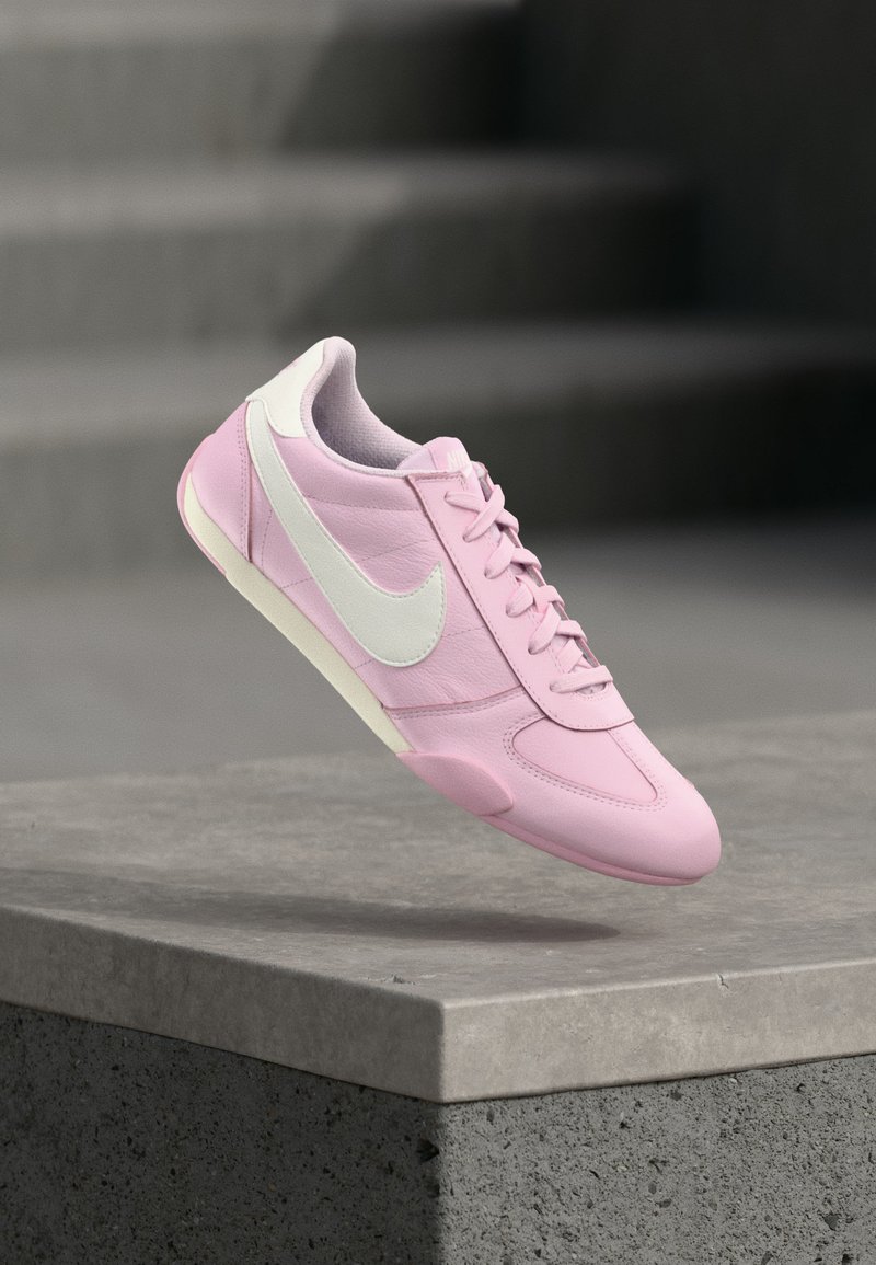 Chaussure de sport rose avec logo swoosh blanc, semelle blanche et lacets, flottant au-dessus d'un rebord en béton gris avec un arrière-plan flou.