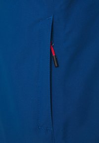 Tejido azul con una textura suave, que presenta un bolsillo lateral y un cordón rojo visible junto con detalles en hardware. La costura es ordenada y consistente.