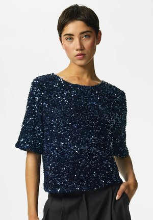 Femme aux cheveux courts bruns portant un haut bleu marine à sequins à manches courtes et un pantalon noir, une main dans la poche.