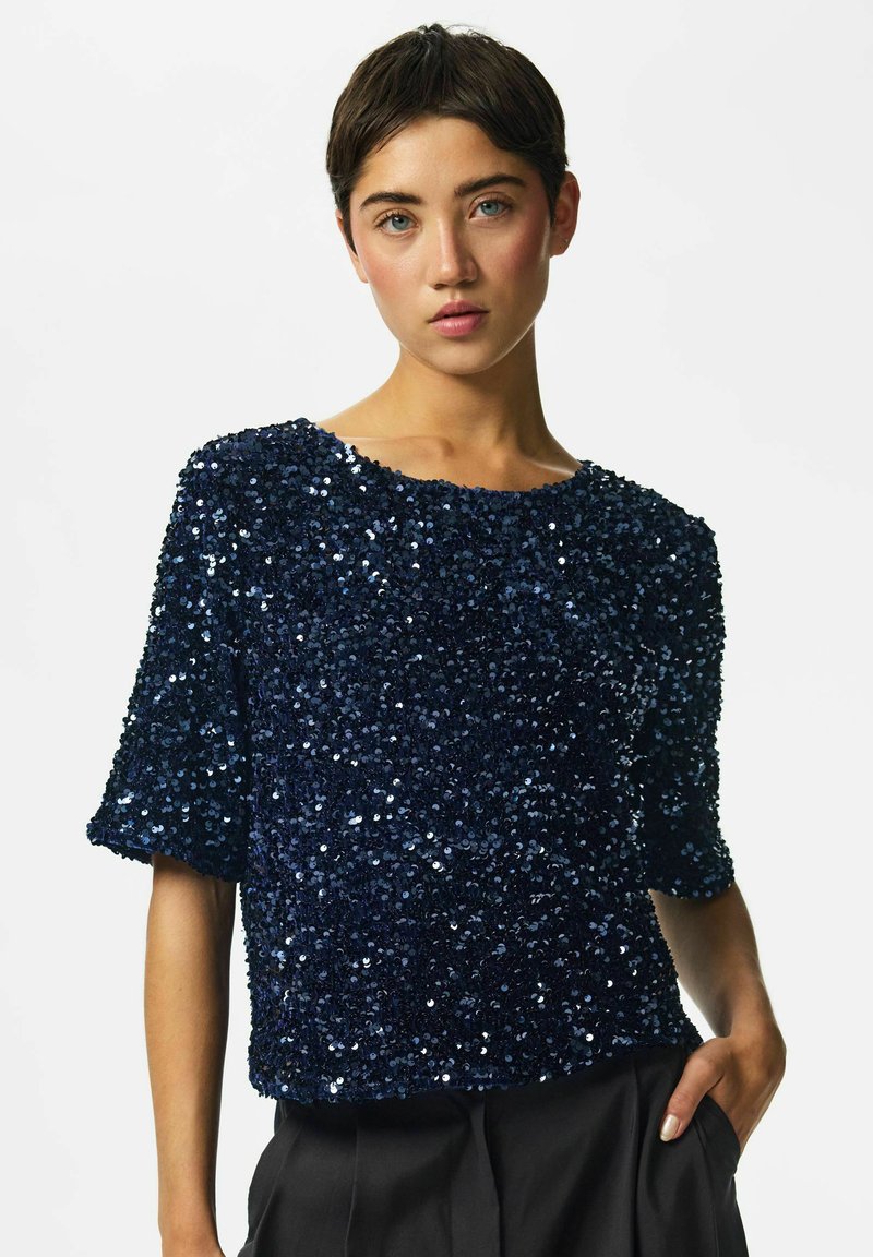 Femme aux cheveux courts bruns portant un haut bleu marine à sequins à manches courtes et un pantalon noir, une main dans la poche.