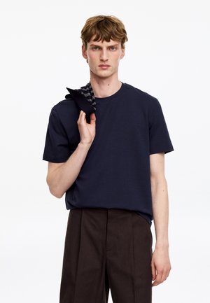 Jeune homme aux cheveux châtain clair portant un t-shirt bleu marine et un pantalon marron foncé, tenant un vêtement rayé plié sur son épaule.
