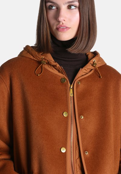 Femme aux cheveux bruns lisses portant un manteau marron à capuche et un col roulé noir, regardant légèrement sur le côté.