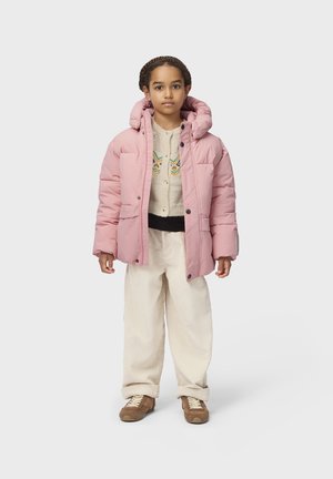 Barn står iført en lyserød puffjakke, lyse mønstret sweater, cremefarvede kordbukser og brune sneakers mod en ensfarvet baggrund.