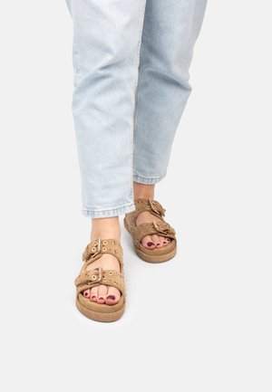 Sandales en daim marron avec double boucle, bout rond et accents d'œillets proéminents. Portées avec un jean clair à revers. Surface lisse.