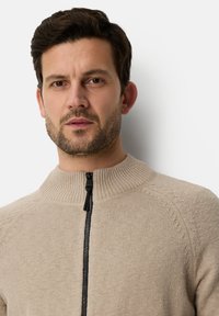 Beige zip-up sweater with ribbed collar and textured knit detail. Features a black zipper and subtle stitch accents on shoulders.