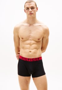 Shorts de boxe noirs avec une ceinture rouge portant le texte "HILFIGER". Le tissu est lisse, ajusté et conçu pour le confort.
