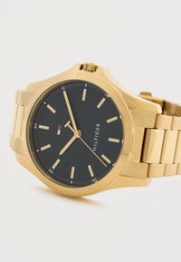 Gold-tone metal wristwatch with black dial, gold hour markers, three hands, and a small red, white, and blue flag emblem near 11 o'clock.