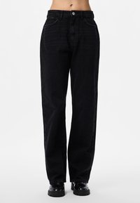 Kvinne iført svarte jeans med høyt liv og rette ben, og svarte chunky loafers, stående mot en ensfarget hvit bakgrunn.