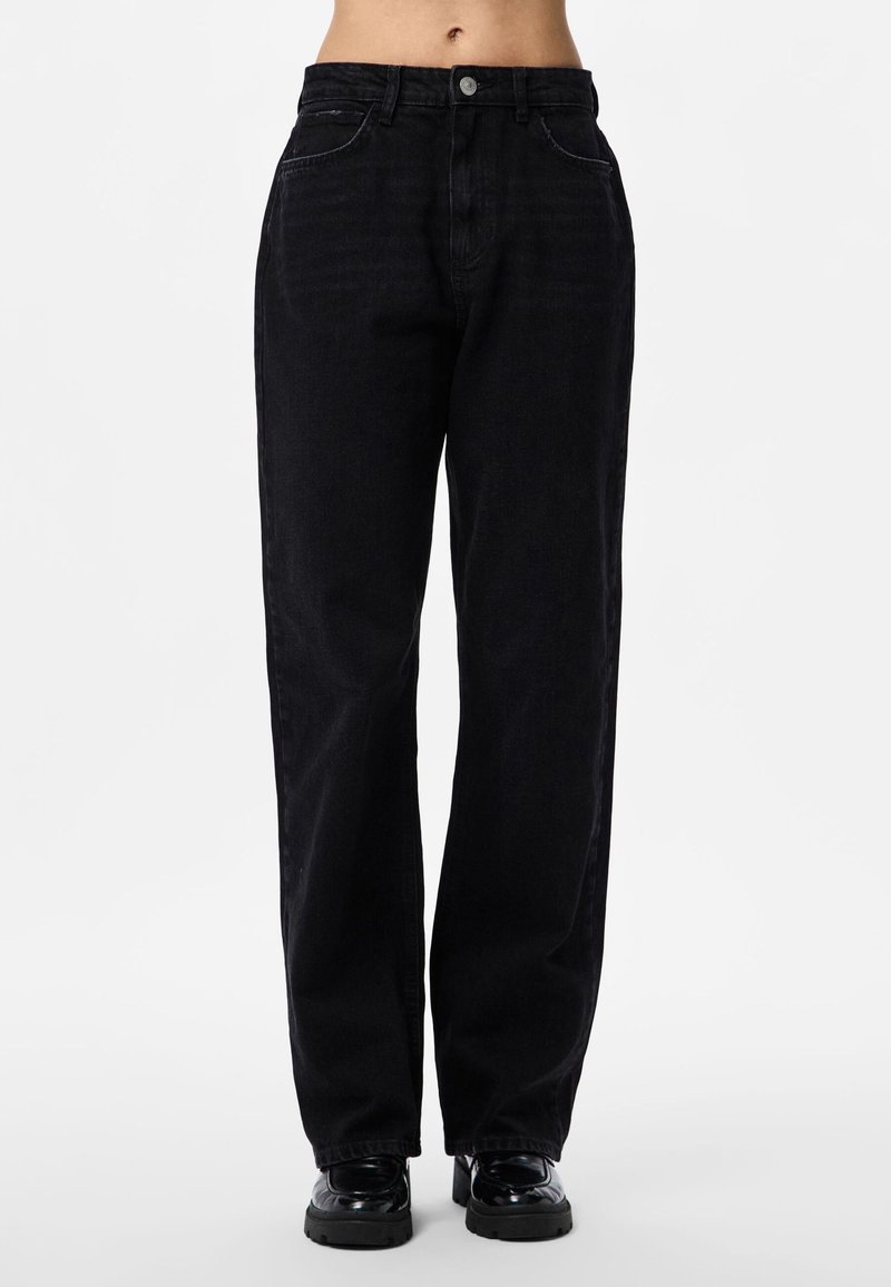 Kvinne iført svarte jeans med høyt liv og rette ben, og svarte chunky loafers, stående mot en ensfarget hvit bakgrunn.