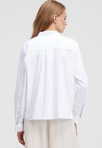 White, long-sleeve shirt with a relaxed fit, a back yoke, and light blue accents on the cuff slits. Soft, lightweight fabric texture.
