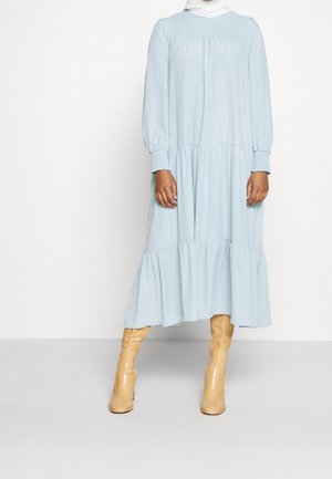 Lichtblauwe, gelaagde jurk met lange mouwen en gerimpelde details, gedragen met beige kniehoge laarzen; de stof lijkt zacht en vloeiend.