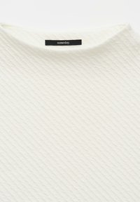 Haut blanc texturé présentant un motif diagonal subtil. L'encolure est arrondie, et une étiquette noire avec du texte blanc est visible.