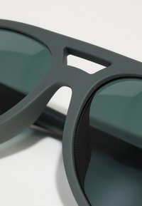 Ochelari de soare gri cu finisaj mat. Au lentile cu nuanță verde și un design unic cu decupaje în ramă pentru ventilație.