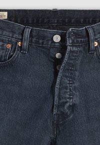 Jeans de ganga azul escuro com cintura alta, com botões de cobre e prata visíveis e um tecido suave e texturizado.