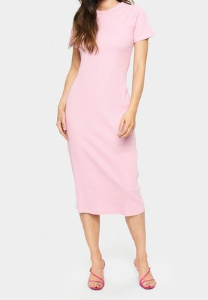 Vrouw draagt een nauwsluitende, lichtroze midi-jurk met korte mouwen, gecombineerd met felroze hoge hakken met bandjes, staand op een effen vloer.