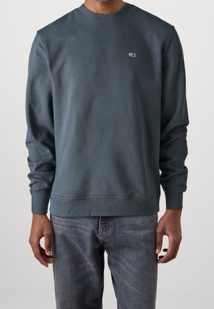 Man draagt een donkergrijze trui met een kleine rechthoekige logo op de linkerborst, gecombineerd met vervaagde zwarte jeans.