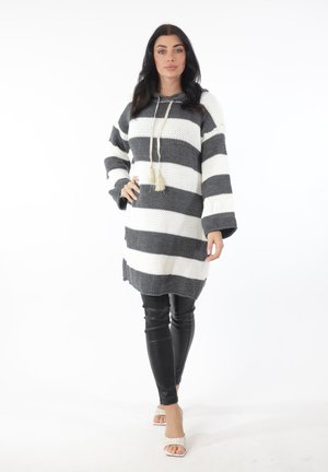 Frau mit langen dunklen Haaren, die ein grau-weiß gestreiftes gestricktes Hoodie-Kleid, schwarze Lederleggings und weiße Sandalen mit Absatz trägt, steht mit einer Hand in der Hüfte.