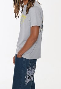 Grijze katoenen T-shirt met korte mouwen, voorzien van gele grafische tekst. Gecombineerd met blauwe spijkerbroeken met een ontwerp van een schedel en vlammen.