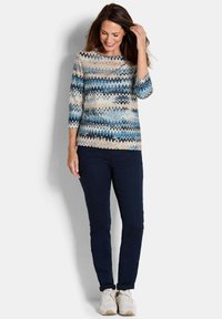 Leichtes, gemustertes Oberteil mit blau-beigefarbenem Zickzackdesign und 3/4-Ärmeln. Kombiniert mit dunklen Jeans und hellen Sneakers.