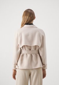 Vaaleanpunainen villasekoitejakku, jossa on vyö, vyötärönauhalla varustetut hihat ja kerroksellinen takamallinen. Päällä mustan paidan ja kermaisten puuvillafarkkujen kanssa.