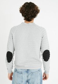 Suéter gris de punto con un patrón texturizado, parches negros en los codos y puños acanalados. Vista trasera; combinado con jeans de mezclilla azul claro.