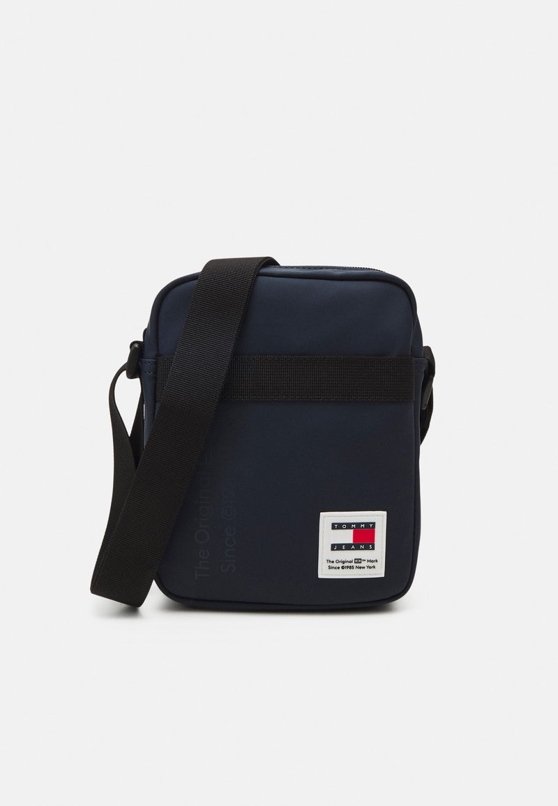 Borsa a tracolla blu con forma rettangolare, realizzata in materiale liscio, dotata di una tracolla nera e un'etichetta bianca con dettagli rossi, blu e neri.