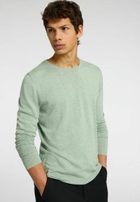 Ljusgrön långärmad skjorta i mjukt tyg med rund hals och åtsittande ärmar, bärs tillsammans med svarta byxor.