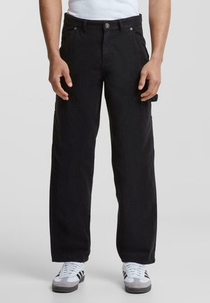 Homme portant un pantalon noir ample et des baskets blanches avec des rayures noires, debout devant un fond gris clair uni.