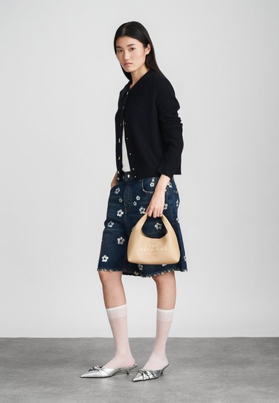 Femme portant un cardigan noir, un haut blanc, une jupe en denim à fleurs, des chaussettes blanches transparentes, des chaussures argentées à bout pointu, tenant un sac beige étiqueté "The Sack Bag."