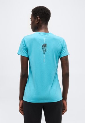 Person med kort krøllet hår iført en lyseblå t-shirt med "The North Face"-logo og tekst på øverste del af ryggen, stående mod en ensfarvet baggrund.