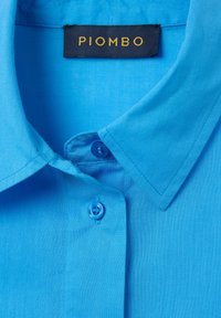 Camisa azul claro con una textura suave, que presenta un collar clásico y dos botones a juego; la etiqueta dice "PIOMBO" en dorado sobre un fondo oscuro.