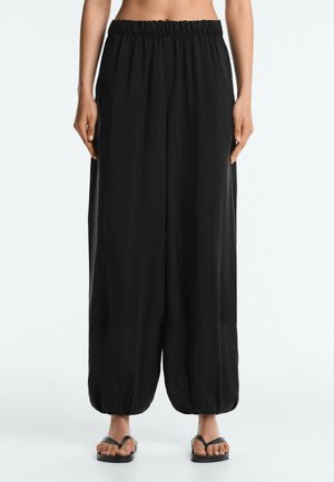 Femme portant un pantalon noir ample à taille élastique avec des poignets froncés et des sandales plates noires, debout devant un fond clair uni.