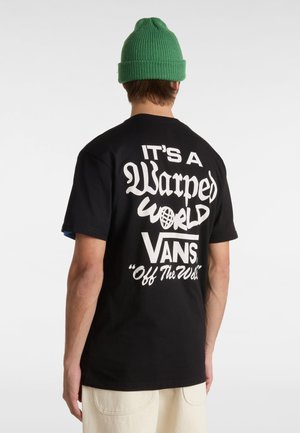 Camiseta de algodón negra con gran texto blanco en la parte de atrás: "ES UN MUNDO Deformado VANS 'Off The Wall'." Acompañada de un gorro de punto verde.