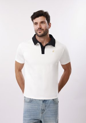 Άνδρας με λευκό πόλο πουκάμισο με μαύρο γιακά και ανοιχτόχρωμα μπλε τζιν, στέκεται με τα χέρια πίσω από την πλάτη σε λευκό φόντο.