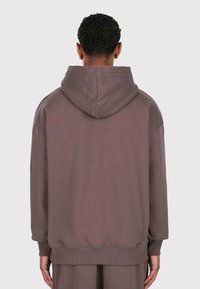 Persona con capelli ricci corti che indossa una felpa oversize marrone semplice e pantaloni abbinati, vista da dietro su uno sfondo bianco.