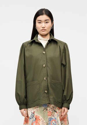 Femme aux cheveux lisses et foncés portant une veste boutonnée vert olive avec de grandes poches frontales, sur une jupe à motif floral.