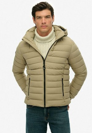 Hombre vistiendo una chaqueta acolchada beige con capucha, un suéter de cuello alto crema y jeans azules, de pie con las manos detrás de la espalda.
