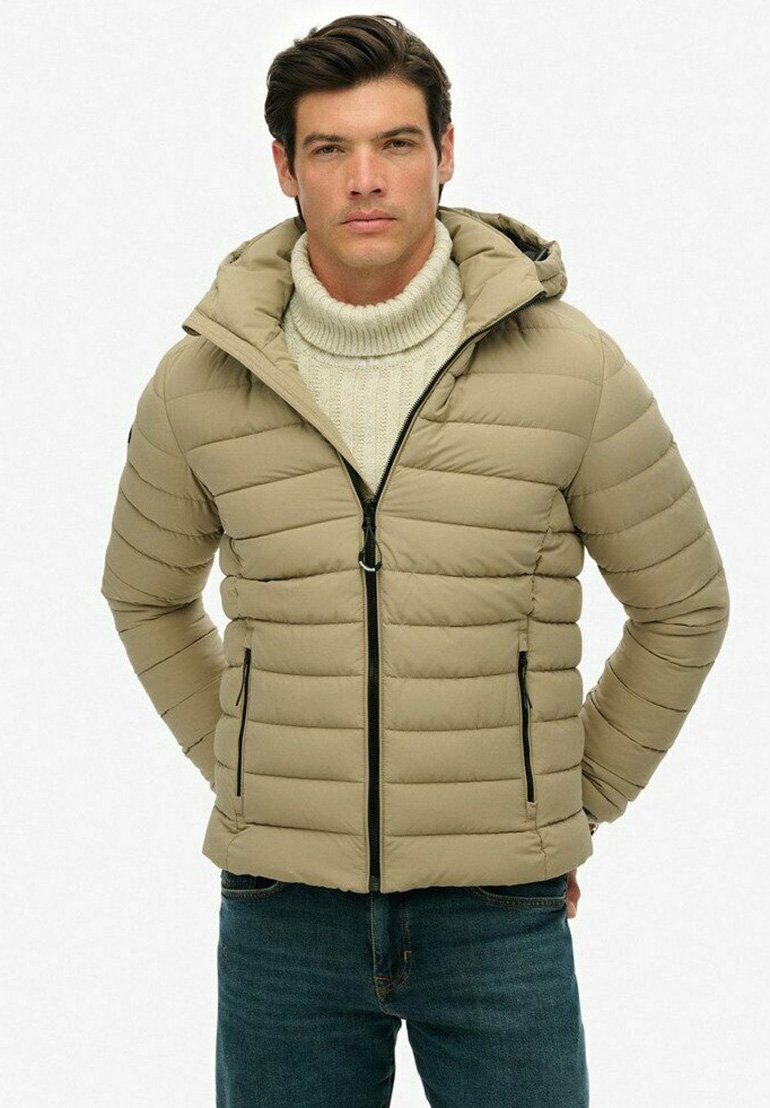 Uomo che indossa un giubbotto trapuntato beige con cappuccio, un maglione a collo alto crema e jeans blue, in piedi con le mani dietro la schiena.