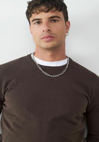 Brauner Strickpullover mit Rundhalsausschnitt, getragen über einem weißen T-Shirt. Besitzt eine silberne Halskette. Glatte Textur, enganliegendes Design.