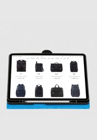 Tablet zeigt einen Onlineshop mit verschiedenen schwarzen Taschen. Die Taschen haben unterschiedliche Designs, Größen und Reißverschlussdetails. Blaue Buchmappe sichtbar.