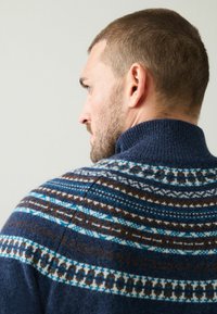 Marineblauer Strickpullover mit hohem Kragen, versehen mit einem geometrischen Muster in Blau, Braun und Weiß über den Schultern und dem oberen Rücken.