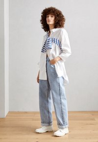 Camisa blanca de gran tamaño con botones y mangas largas, detalle anudado de rayas azules y blancas, jeans de cintura alta en azul claro y zapatos blancos.