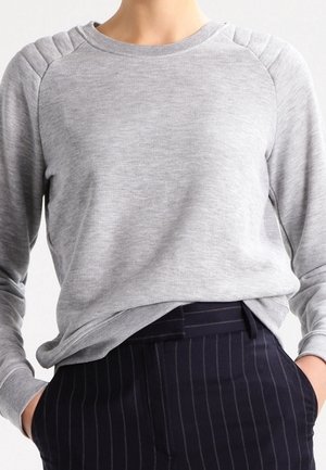 Personne portant un sweat-shirt gris clair côtelé avec des épaules rembourrées et un pantalon bleu marine à fines rayures avec les mains dans les poches.