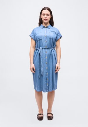 Femme debout, de face, portant une robe bleu à manches courtes boutonnée jusqu'au genou avec une ceinture nouée à la taille et des chaussures noires.