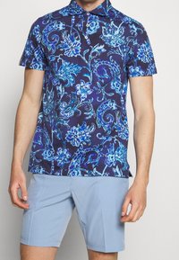 Pánská polokošile s krátkým rukávem v námořnické modré barvě s modrým květinovým paisley potiskem, se límečkem a knoflíkovou légou na tři knoflíky, v kombinaci se světle modrými šortkami.