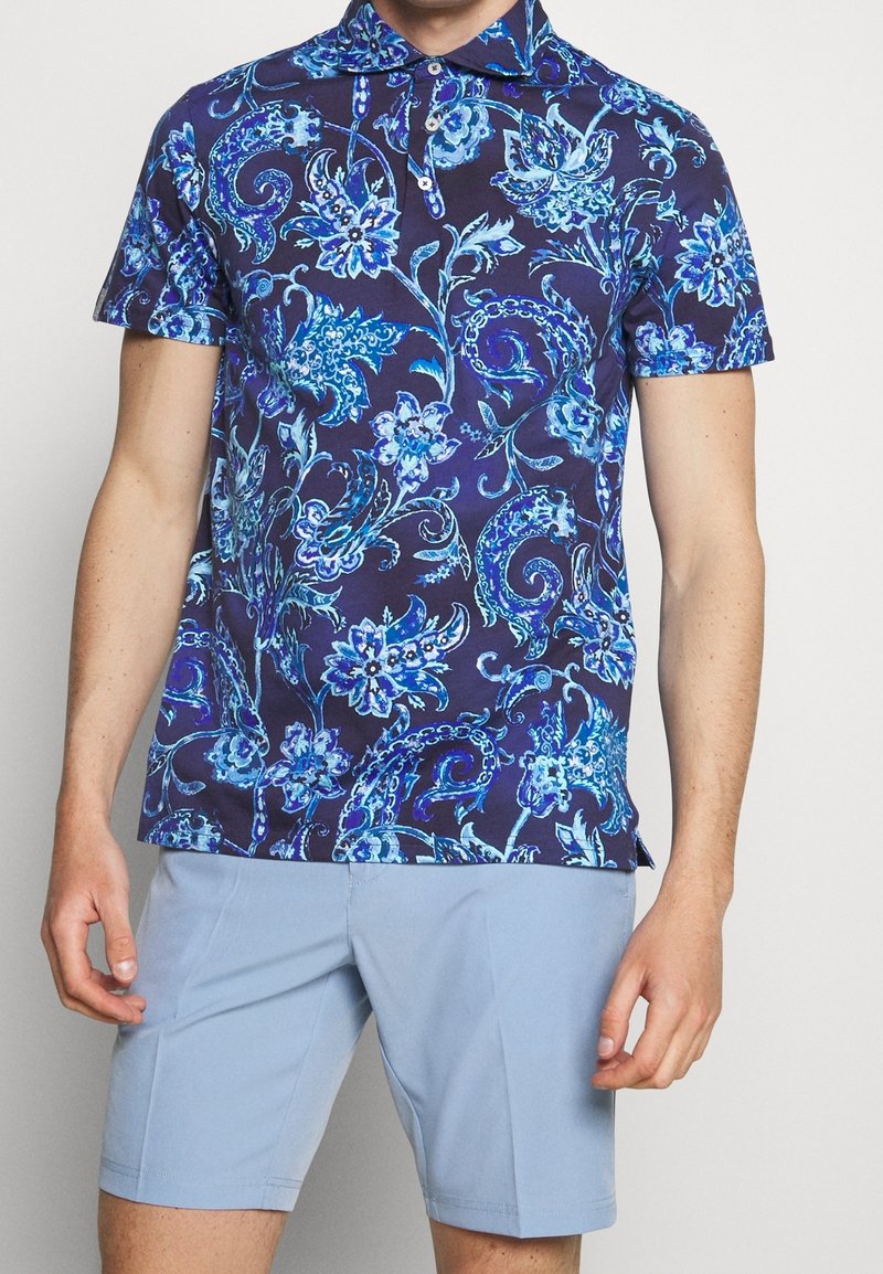Pánská polokošile s krátkým rukávem v námořnické modré barvě s modrým květinovým paisley potiskem, se límečkem a knoflíkovou légou na tři knoflíky, v kombinaci se světle modrými šortkami.