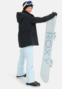 Snowboardzista w czarnej kurtce, jasnoniebieskich spodniach i czarnych butach trzyma szary snowboard z dużymi, jasnoniebieskimi literami "ROXY".